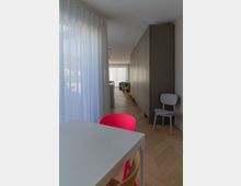 Wohnraum in modernem Stil mit hellem Holzboden und großen weißen Vorhängen, die Tageslicht durchlassen. Im Vordergrund steht ein weißer Tisch mit einem pinkfarbenen und einem hellen Holzstuhl, während im Hintergrund ein Sofa und grüne Pflanzen sichtbar sind.