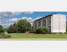 Ein mehrstöckiges Wohngebäude mit Balkonen, umgeben von Bäumen, Büschen und einer großen Grünfläche. Im Vordergrund verläuft ein schmaler Weg, und links im Bild steht ein Strommast.