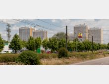 Mehrere hohe Wohnblöcke im Hintergrund, umgeben von Bäumen entlang einer Straße. Im Vordergrund verlaufen Bahngleise und Gräser, rechts ist ein McDonald's-Schild sichtbar.