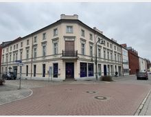 Eckgebäude mit hellbeiger Fassade im klassischen Stil, verziert mit Stuckdetails und großen Fenstern, an einer gepflasterten Straßenecke. Daneben stehen moderne Gebäude mit roten Klinker- und Glasfassaden; im Vordergrund Gehwege, Straßenschilder und parkende Autos.