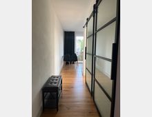 Schmaler, moderner Flur mit hellem Holzfußboden; rechts eine Schiebetür mit Milchglas und schwarzem Rahmen, links eine gepolsterte Bank. Im Hintergrund stehen zwei schwarze Stühle vor einem Fenster mit Blick ins Freie, verdeckt von einem dunklen Vorhang.