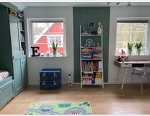 Kinderzimmer mit Holzboden, Spielteppich und zwei Fenstern. Links ein eingebautes grünes Regal mit Schlafplatz, in der Mitte ein blaues Truhenmöbel und ein Regal voller Bücher und Spiele, rechts ein Schreibtisch mit Pflanzen auf der Fensterbank. Im Hintergrund ist ein rotes Haus sichtbar.