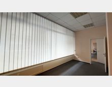 Büro- oder Geschäftsräume mit beigen Wänden und grauem Teppichboden, großen Fenstern mit weißen Vertikaljalousien, einer Heizkörperleiste unter den Fenstern und einer Tür, die in einen angrenzenden Raum führt. Die Decke ist mit einem Raster aus abgehängten Deckenplatten und eingebauten Leuchten versehen.