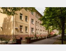 Wohngebäude aus der Nachkriegszeit mit drei Etagen, gelb- und rosafarbenen Putzfassaden, einem Sockel aus rotem Backstein sowie doppelt verglasten Fenstern; davor eine baumbestandene Gehweg-Promenade mit niedrigen roten Backsteinmauern und schattenspendenden Bäumen.