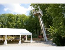 Ein Biergarten mit Reihen von Holzsitzgarnituren unter einem weißen Zelt vor einer Kulisse aus hohen, grünen Bäumen. Rechts steht ein schräger, rostiger Stahlaufbau mit einer Aussichtsplattform.