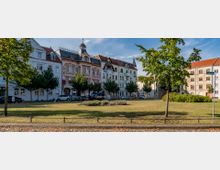 Offener Stadtplatz mit einer Grünfläche im Vordergrund, umgeben von Altbauten, darunter ein verziertes, rosa-weißes Gebäude mit einem Turm. Gepflegte Bäume säumen den Platz, und Autos sind entlang der Gebäude geparkt.