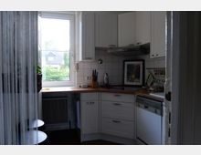 Kleine, helle Küche mit weißen Einbauschränken und Holzarbeitsflächen. Ein Fenster mit Blick ins Grüne bringt Tageslicht, daneben stehen Küchenutensilien und ein Messerblock; rechts befindet sich eine Spülmaschine.