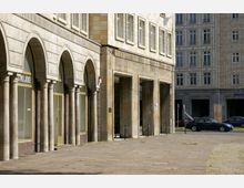 Bogenförmige Arkaden mit massiven Steinsäulen prägen die Fassade eines Gebäudes mit gelblich beigen Kacheln und rechteckigen Fenstern. Im Hintergrund stehen weitere Gebäude in ähnlichem Baustil sowie parkende Autos am Straßenrand.