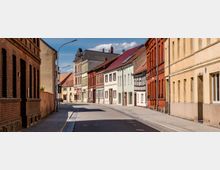 Straßenszene mit historischen Wohngebäuden in verschiedenen Farben, darunter gelbe, rote und pastellfarbene Fassaden, einige im Fachwerkstil; die Straße ist gepflastert und von Bürgersteigen gesäumt, bei sonnigem Wetter und blauem Himmel.