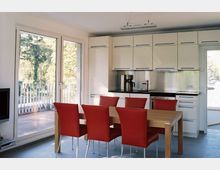 Moderne Küche mit weißen Einbauschränken, silberfarbener Rückwand und schwarzer Arbeitsfläche. Im Vordergrund steht ein Holzesstisch mit sechs roten Stühlen; links eine große Balkontür mit Blick auf grüne Bäume.