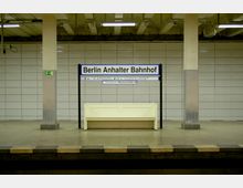 U-Bahn-Station mit grauen Fliesenwänden, zwei Pfeilern und einer Sitzbank aus hellem Material. Ein Schild mit der Aufschrift "Berlin Anhalter Bahnhof" befindet sich zentral über der Bank.