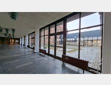 Langer, überdachter Gebäudeflur mit dunklem Steinboden und einer durchgehenden Reihe großer, schwarz gerahmter Glasfenster und -türen. Durch die Fenster ist ein gepflasterter Innenhof mit niedrigen Gebäuden und einer Backsteinfassade zu sehen.