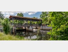 Überdachte Brücke mit metallischer Struktur über einem Fluss, umgeben von grünen Bäumen und Gras; im Vordergrund ein ruhiger Wasserlauf mit leichten Stromschnellen unter der Brücke.