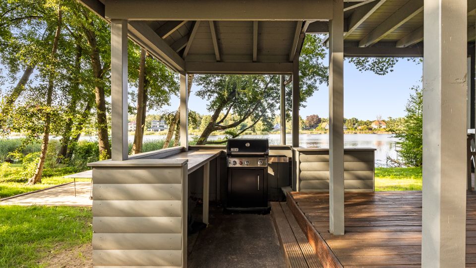 Überdachter Grillplatz aus Holz mit eingebautem Gasgrill und umlaufender Arbeitsfläche auf einer Terrasse. Im Hintergrund liegen Wiese, Bäume und ein Seeufer mit Blick auf Häuser am gegenüberliegenden Ufer.