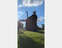 Hohe, dunkle Holzwindmühle mit großen Flügeln auf einer offenen Wiese, aufgenommen vor blauem Himmel mit Wolken. Im Vordergrund stehen Gras, ein kleiner Baum und einfache Sitzgelegenheiten; im Hintergrund sind weitere kleine Gebäude und Zäune zu sehen.
