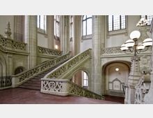 Großes Treppenhaus in einem historischen Gebäude mit kunstvoll verzierten Steinbalustraden, hohen Rundbogenfenstern, Säulen und einem mehrarmigen Kronleuchter mit Kugellampen. Die Wände sind hell, und der Boden besteht aus rotem Stein.