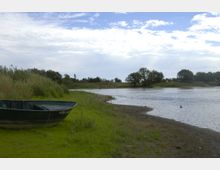 Ein Flussufer mit flachem, sandigem Boden und grünem Grasbewuchs. Im Vordergrund liegt ein grünes Ruderboot am Ufer, im Hintergrund sind Bäume und Büsche unter einem teilweise bewölkten Himmel zu sehen.