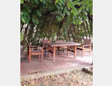 Holztisch mit vier passenden Holzstühlen auf einer gepflasterten Fläche, umgeben von dichtem, grünem Blattwerk, das Schatten spendet. Die Umgebung wirkt wie ein Garten oder eine Terrasse.