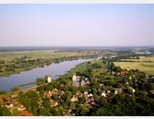 Luftaufnahme einer ländlichen Umgebung mit einem Fluss, der sich durch grüne Wiesen und Bäume schlängelt. Im Vordergrund ist ein kleines Dorf mit Wohnhäusern, einzelnen Gebäuden und zwei markanten weißen Türmen zu sehen.
