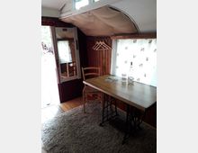 Kleiner Innenraum mit Holzwänden, einem Holztisch auf einem Metallgestell, einem Holzstuhl und einem Teppichboden. An der Wand befinden sich mehrere Holzbügel, und ein Fenster mit weißem Vorhang lässt Licht herein.