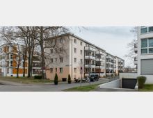 Wohngebiet mit mehrstöckigen Wohngebäuden, die helle Fassaden und Balkone besitzen, umgeben von Bäumen und kleinen Grünflächen. Im Vordergrund sind eine kleine Einfahrt zur Tiefgarage, parkende Fahrzeuge und Gehwege zu sehen.