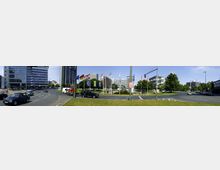 Eine städtische Kreuzung mit mehreren Fahrbahnen, umgeben von modernen Bürogebäuden und Hochhäusern. In der Mitte des Kreisverkehrs stehen Fahnenmasten mit verschiedenen Nationalflaggen, und eine gepflegte Grünfläche mit Bäumen ergänzt die urbane Umgebung.