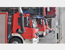 Innenansicht einer Feuerwache mit mehreren geparkten roten Feuerwehrfahrzeugen in einer Reihe. An den Fahrzeugen sind unter anderem große Abgasrohre angeschlossen, und die Umgebung besteht aus grauen Bodenfliesen und Betonwänden.