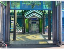 Eingangsbereich einer modernen Bahnhaltestelle mit farbigen Glaswänden in Blau- und Grüntönen. Im Vordergrund sind senkrechte Metallstützen, Fliesenboden und ein Fahrradkorb zu sehen, im Hintergrund führen Treppen und ein Aufzug nach oben.