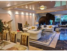 Wohnzimmer mit luxuriösem und dekorativem Design, ausgestattet mit weißen Polstermöbeln, einem großen Teppich mit Blumenmuster und einer auffälligen Elefantenstatue. Im Raum befinden sich weitere dekorative Elemente wie eine Engelsfigur, goldene Akzente an Möbeln und ein großer Fensterbereich mit Blick auf einen grünen Garten.