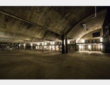 Weitläufiger, industrieller Innenraum mit hohen Betonbögen und einer geschwungenen Decke. Wände und Boden wirken robust, während warme Beleuchtung die Struktur aus Pfeilern und großen Fenstern betont.