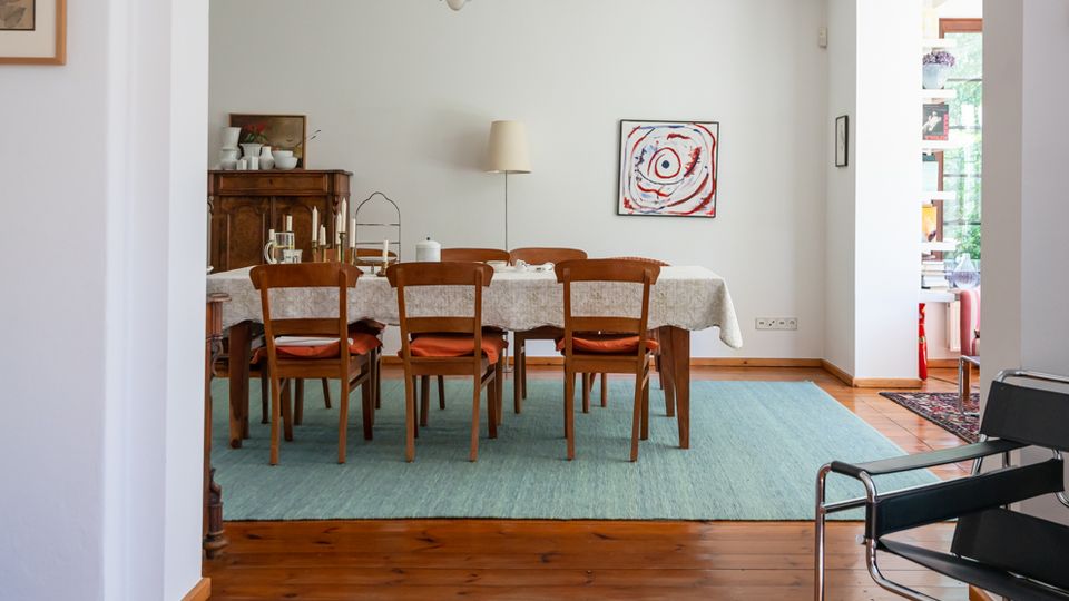 Heller Essbereich in einem Wohnhaus mit langem Holztisch und sechs Stühlen auf einem großen blauen Teppich, Holzdielenboden und einer Deckenleuchte. An der Wand hängen ein abstraktes Bild, daneben steht ein Sideboard; rechts öffnet sich der Raum zu einem angrenzenden Bereich mit Regal und Fenster.