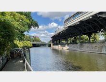 Ein Fluss mit befestigten Ufern, daneben ein schmaler Fußweg mit Geländer und Bäumen. Rechts erhebt sich eine hohe Eisenbahnbrücke mit Stahlträgern, im Hintergrund eine Straßenbrücke und Gebäude.