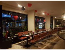 Restaurant-Innenraum mit modernem Design: Eine lange Tafel ist mit braunen Lederstühlen und gedeckten Plätzen arrangiert, dekoriert mit Herzluftballons und hängenden Ornamenten. Große Fensterfronten zeigen eine nächtliche Straßenszene mit Lichtern und vorbeifahrenden Fahrzeugen.