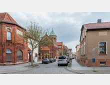 Straßenszene in einem Wohnviertel mit Kopfsteinpflaster, links ein historisches Gebäude aus rotem Backstein mit dekorativen Bögen, im Hintergrund eine Kirche mit grünem Turmdach. Rechts stehen moderne Wohnhäuser mit Putzfassaden, und geparkte Autos säumen die Straße.