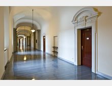 Ein langer, heller Flur mit glänzendem Boden und gewölbter Decke, gesäumt von Holzfenstern und ornamentalen Türrahmen. An den Wänden hängen antik wirkende Lampen, die den Korridor beleuchten.