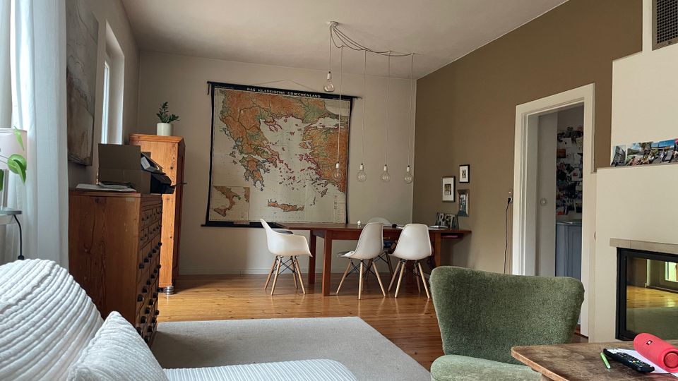 Wohn- und Essbereich mit Holzboden, großem Wandkarten-Poster und Esstisch mit vier weißen Stühlen unter hängenden Glühbirnen. Links stehen eine Kommode und ein Sofa am Fenster, rechts sind ein Durchgang zu einem weiteren Raum sowie ein Kamin und ein Sessel zu sehen.