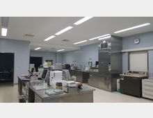 Ein moderner Laborraum mit hellen Wänden und Deckenbeleuchtung. Der Raum ist mit Arbeitsflächen, Laborgeräten, Kühlschränken aus Edelstahl und Aufbewahrungsschubladen ausgestattet, wobei diverse chemische und elektronische Geräte sichtbar sind.