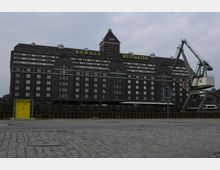 Großes Backsteingebäude mit der Aufschrift „BEHALA WESTHAFEN“, flankiert von einem alten Hafenkran auf der rechten Seite; gepflasterte Hafenfläche und gelbes Eingangstor im Vordergrund.