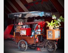 Ein mobiler Kaffeewagen in Form eines Lastenfahrrads mit einer schwarzen Markise, auf der „coffee-bike“ steht. Der Wagen ist mit einer Kaffeemaschine, einem Becherhalter, einem Glasbehälter für Gebäck sowie einer Speisekarte ausgestattet. Daneben stehen Topfpflanzen als Dekoration.