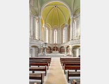 Innenansicht einer historischen Kirche mit hohen Rundbögen und einem hellgrünen, gewölbten Deckengewölbe. Im Vordergrund befinden sich Holzbänke, während der Altarbereich mit einem großen Holzkreuz und einer erhöhten Kanzel im Hintergrund sichtbar ist.