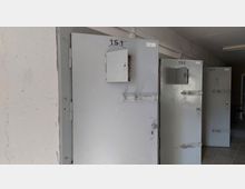 Ein Flur mit mehreren grauen Metalltüren, die jeweils mit Schließmechanismen und kleinen, nach außen öffnenden Klappen versehen sind. Über den Türen befinden sich handgeschriebene Nummern, die die einzelnen Räume kennzeichnen.