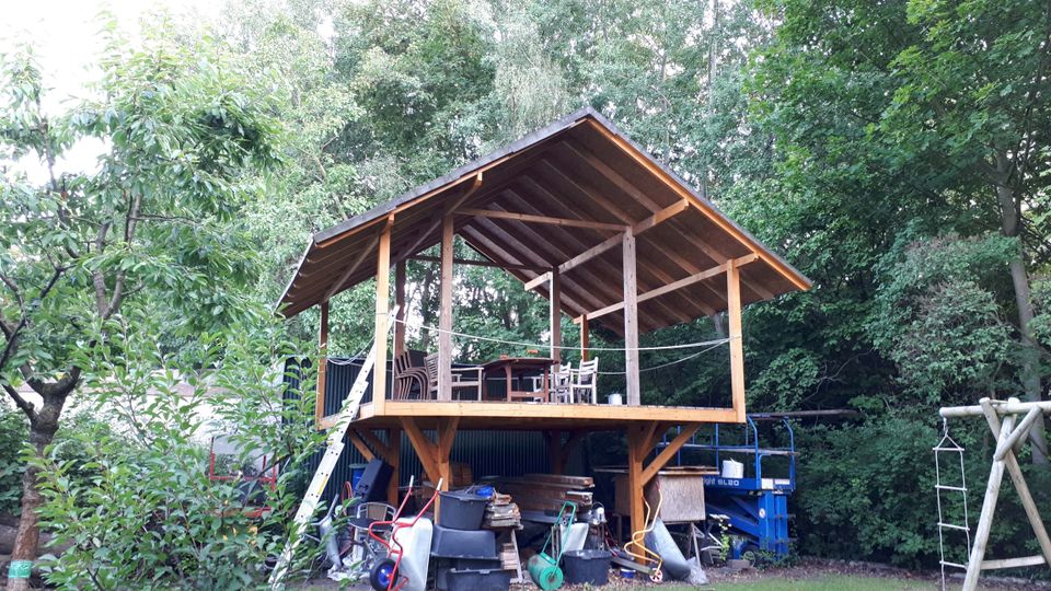 Erhöhte Holzterrasse mit offenem Satteldach und Sitzmöbeln, am Rand einer Rasenfläche vor dichtem Wald. Unter der Plattform stehen gestapelte Materialien und Gartengeräte; seitlich ist eine Leiter angelehnt.