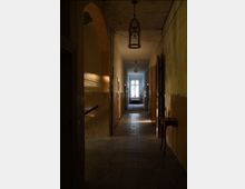 Ein langer, schmaler Korridor in einem alten Gebäude mit abblätternden Wänden, einer abgenutzten Steinbodenfliese und mehreren offenen Türen. Am Ende des Flurs befindet sich ein Fenster, durch das natürliches Licht einfällt, sowie hängende Lampen von der Decke.