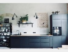 Moderne Küche mit mattschwarzen Schränken und Holzarbeitsplatten, einer schwarzen Spüle und integriertem Backofen. Im Hintergrund sind ein offenes Regal mit Geschirr und Gläsern, ein weiß gestrichener Ziegelabschnitt sowie Dekorationen wie Pflanzen und Wandlampen zu sehen.