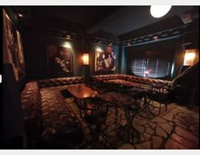 Dunkel beleuchtete Bar mit braunen Ledersofas, runden Holztischen und Metallstühlen. Wände sind mit Bildern dekoriert, und ein Fenster mit Jalousien lässt gedämpftes Licht in den Raum.