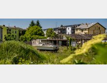 Eine verwilderte Fläche mit einem verlassenen Boot im Vordergrund und einer verfallenen Holzkonstruktion im Hintergrund, umgeben von Gräsern, Büschen und Bäumen; dahinter stehen Wohnhäuser in Pastellfarben und moderner Bauweise.