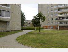 Außenbereich eines Plattenbau-Wohnkomplexes mit mehreren mehrstöckigen Gebäuden aus Beton, Balkonen und Graffiti an den Wänden. Dazwischen befindet sich eine Grünfläche mit Gehwegen, einem kleinen Baum und Büschen.