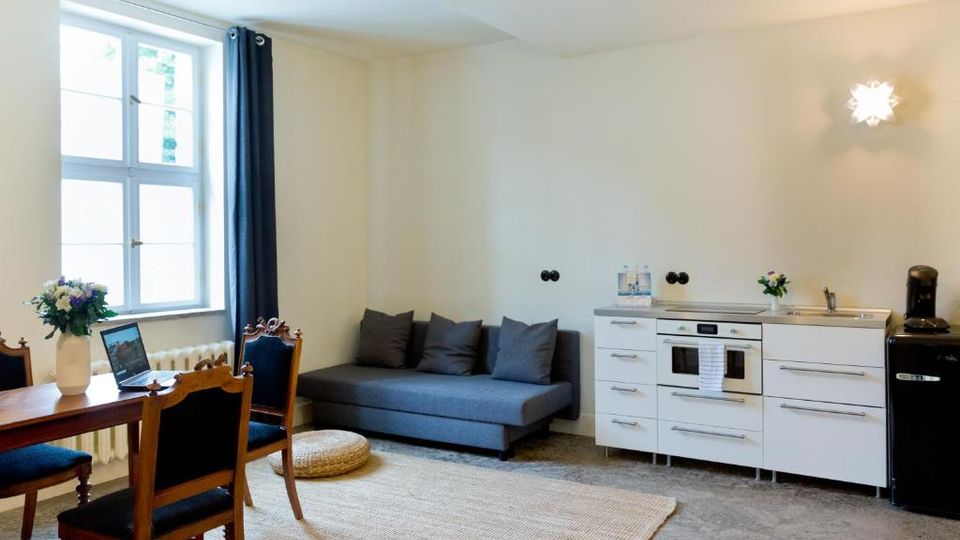 Heller Wohnraum mit offener Küchenzeile: links ein großes Fenster mit dunklem Vorhang sowie ein Esstisch mit Stühlen, mittig ein graues Sofa auf einem Teppich. Rechts eine weiße Küchenzeile mit Spüle und Backofen neben einem schwarzen Kühlschrank, darüber eine Wandleuchte.