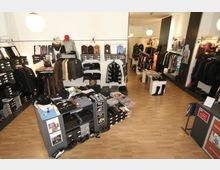 Ein kleiner Modegeschäftsraum mit hölzernem Boden, Regalen voller Kleidung, gestapelter Ware und Schränken für Schuhe. Die Einrichtung besteht aus Schaufensterpuppen, Kleidung auf Bügeln und Tischflächen mit gefalteten Textilien; Lampen an der Decke sorgen für Beleuchtung.