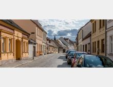 Kopfsteinpflasterstraße in einer ruhigen Wohngegend mit historischen Reihenhäusern in Pastellfarben auf beiden Seiten. Geparkte Autos stehen entlang der Straße, und im Hintergrund ist eine Kirchturmspitze zu sehen.